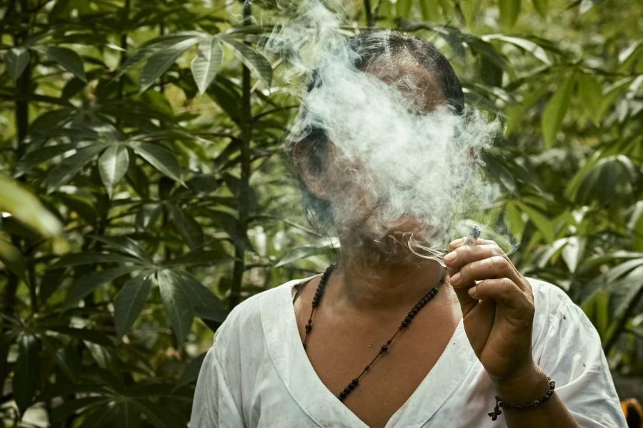 Tobacco is smoked by Nayive, the mother and grandmother of the community, in front of her cassava chagra (cultivation area). Smoke from her mouth protects, cleans, and guards both body and land. Nayive is famous for her great knowledge of plants and ancestral medicine, standing for feminine wisdom that nurtures and brings balance. In her action, the link between the material and the spiritual appears. Like many women in the Amazon, she guards the knowledge and sacred relationship with the earth.